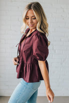 Woman wearing a burgundy blouse with puffed sleeves against a white brick wall.
