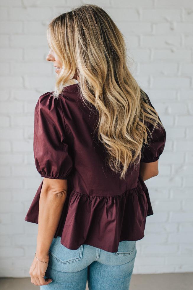 Woman wearing a burgundy blouse with ruffled details and light blue jeans against a white brick wall.