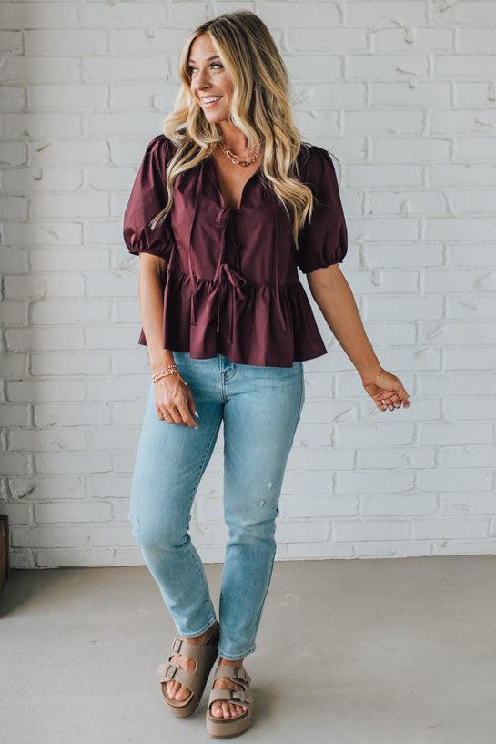 Woman wearing a burgundy blouse and light blue jeans against a white brick wall.