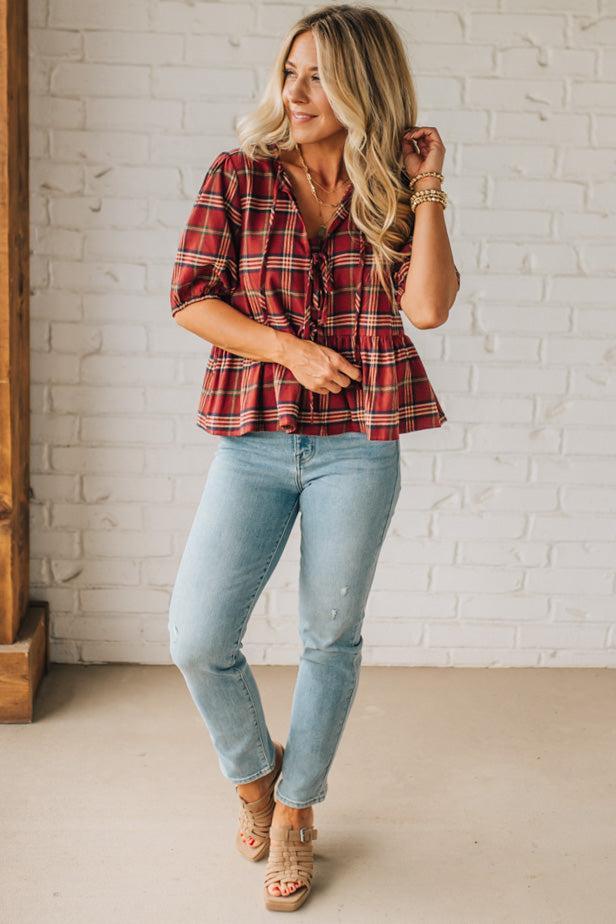 Woman wearing a red plaid shirt and light blue jeans against a white brick wall.