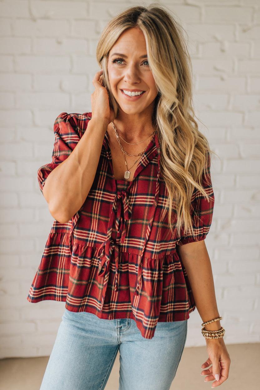 Woman wearing a red plaid top and light blue jeans against a white brick wall.