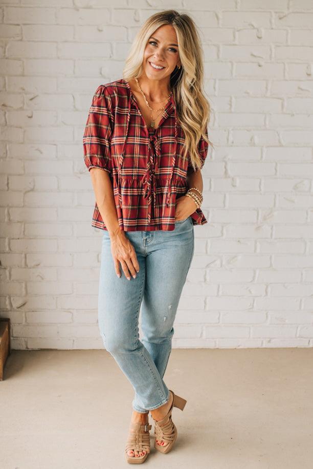 Woman wearing a red plaid shirt and light blue jeans against a white brick wall.