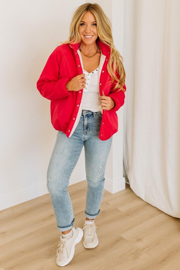 Woman wearing a red jacket, white shirt, blue jeans, and white sneakers indoors.
