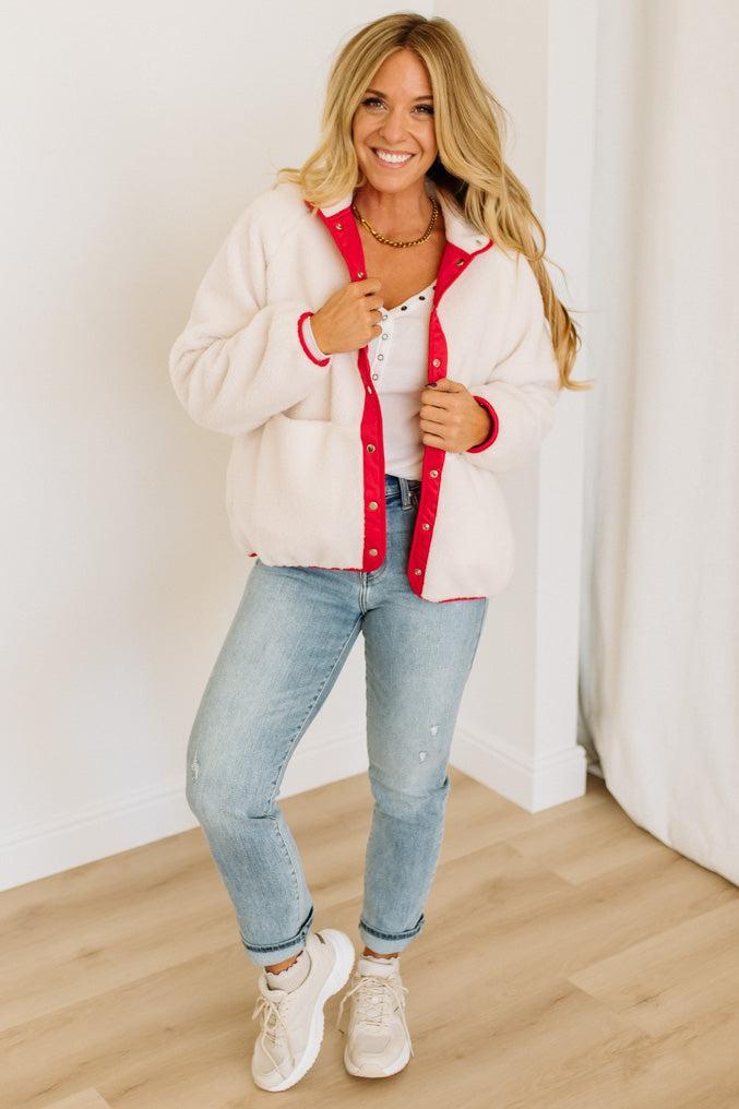 Woman wearing a white jacket with red trim, white shirt, blue jeans, and white sneakers indoors.