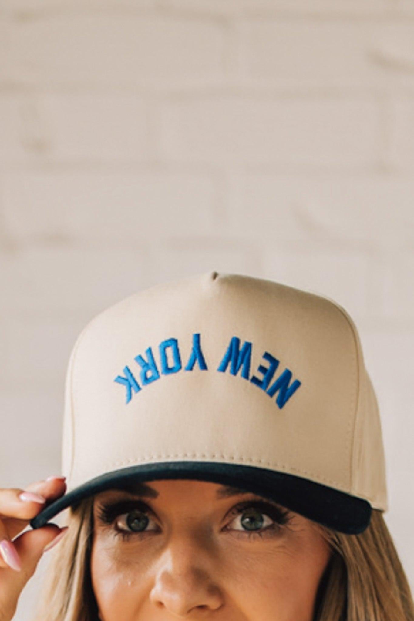 Blonde Woman wearing a Trucker Hat with Blue Upside Down embroidered: New York at the front center. Cream hat with Black Brim and accents.