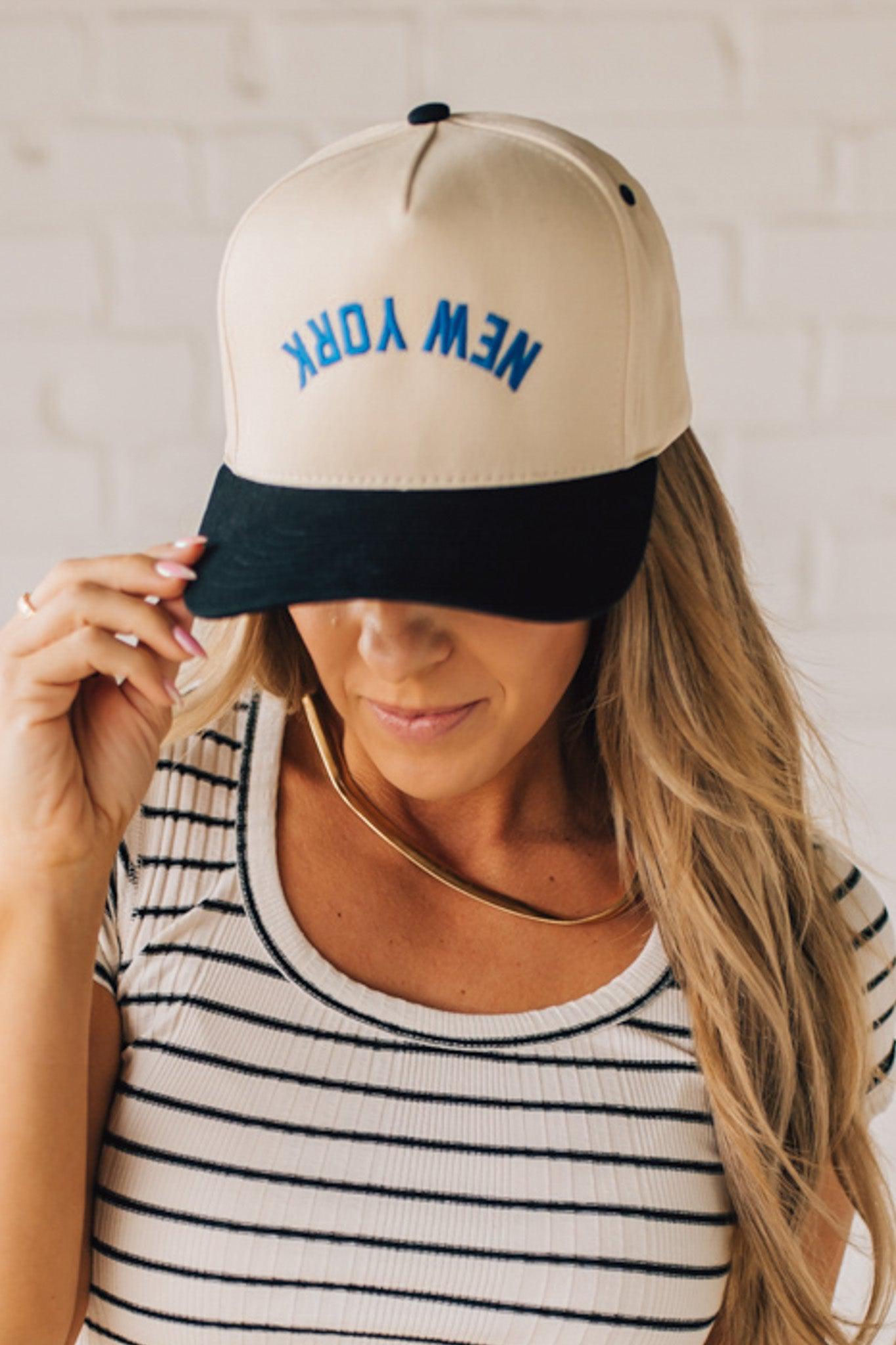 Blonde Woman wearing a Trucker Hat with Blue Upside Down embroidered: New York at the front center. Cream hat with Black Brim and accents.