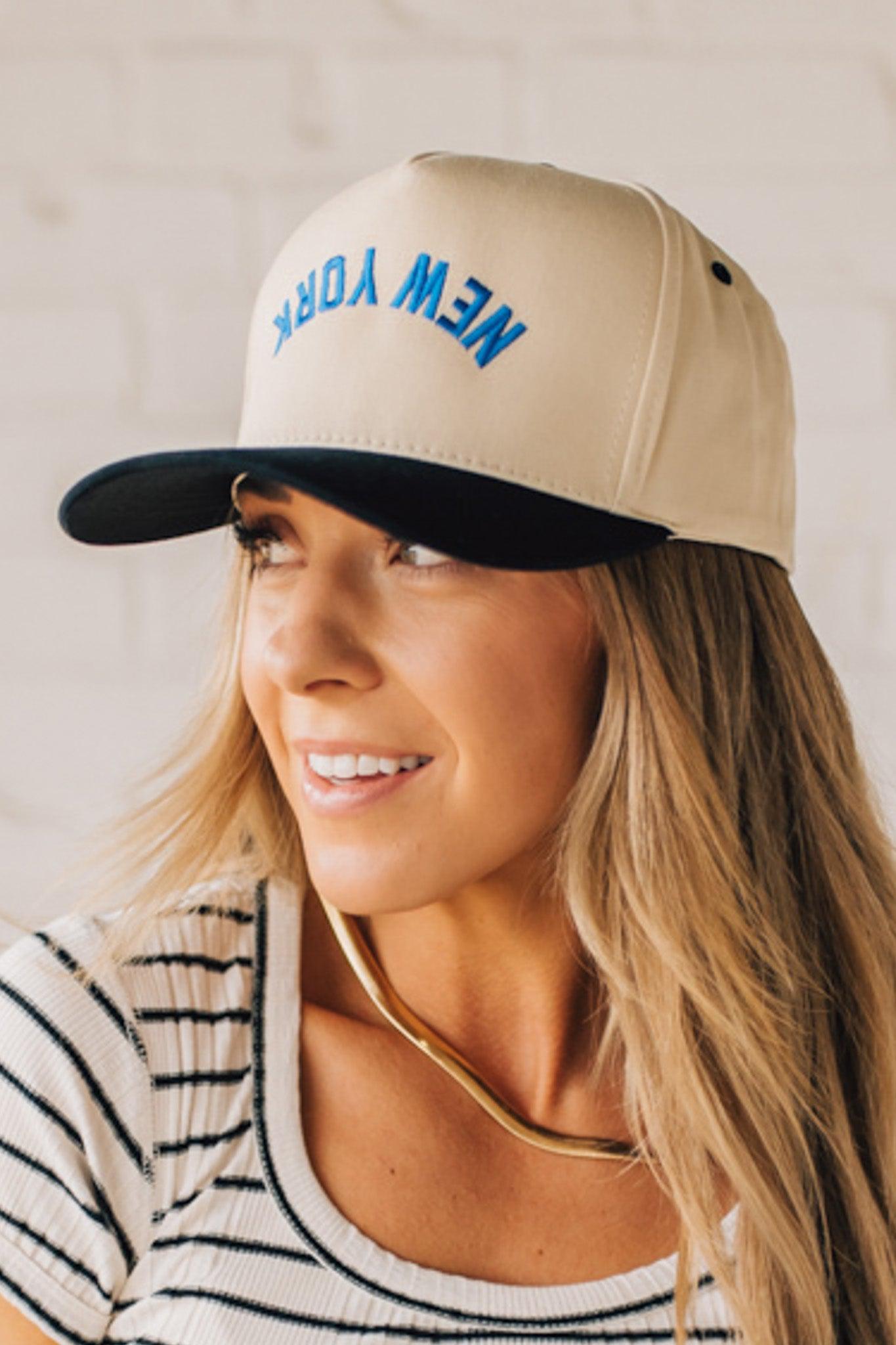 Blonde Woman wearing a Trucker Hat with Blue Upside Down embroidered: New York at the front center. Cream hat with Black Brim and accents.