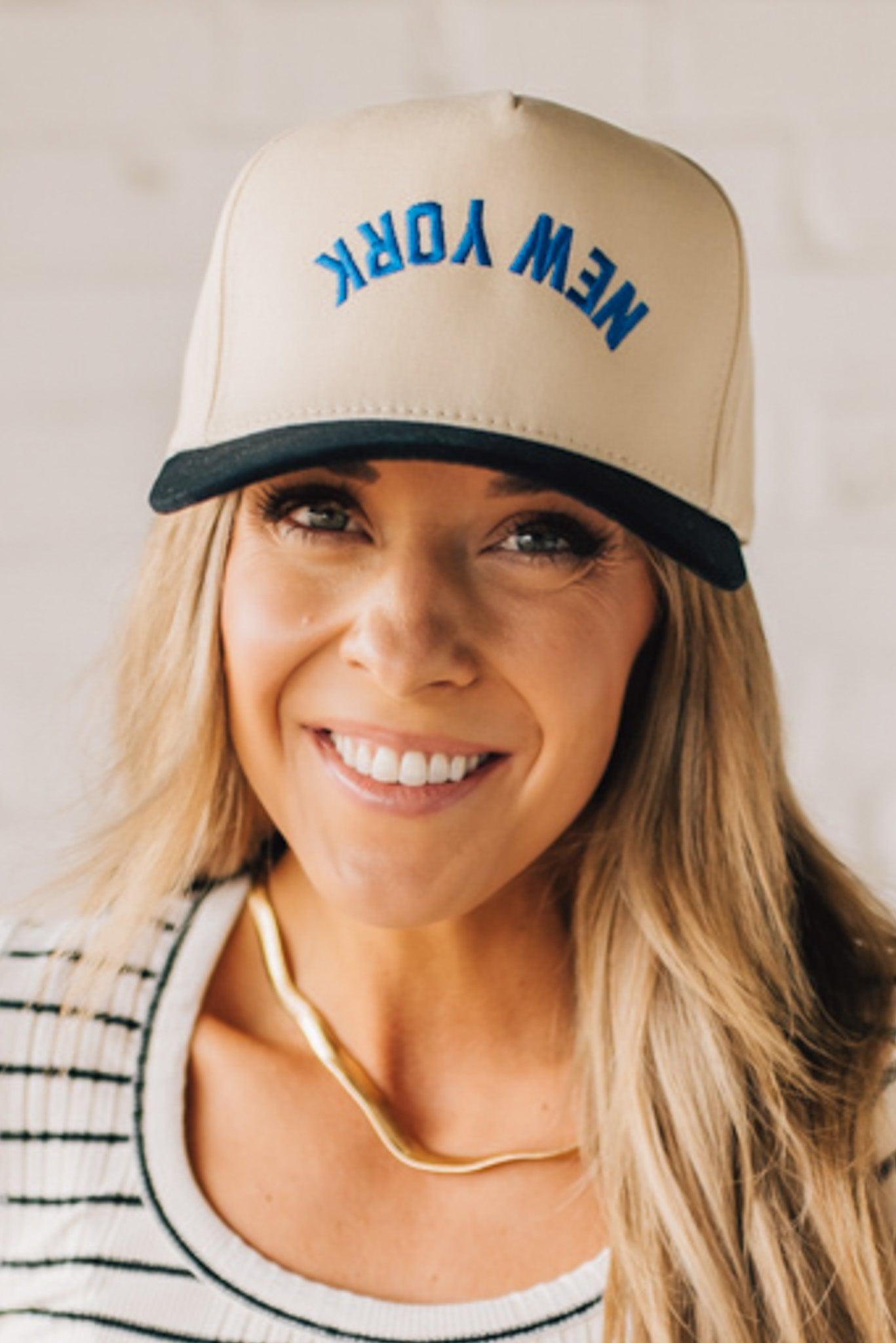 Blonde Woman wearing a Trucker Hat with Blue Upside Down embroidered: New York at the front center. Cream hat with Black Brim and accents.