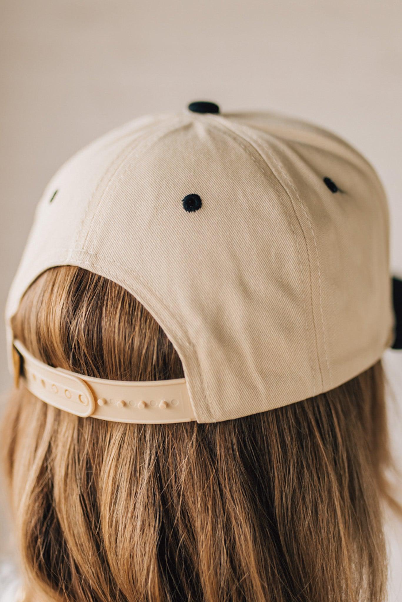 Blonde Woman wearing a Trucker Hat with Blue Upside Down embroidered: New York at the front center. Cream hat with Black Brim and accents.