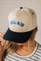 Blonde Woman wearing a Trucker Hat with Blue Upside Down embroidered: New York at the front center. Cream hat with Black Brim and accents.