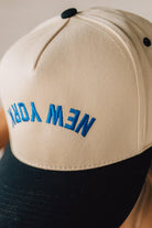 Blonde Woman wearing a Trucker Hat with Blue Upside Down embroidered: New York at the front center. Cream hat with Black Brim and accents.