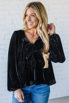 Woman wearing a black velvet blouse with blue jeans against a white brick wall.