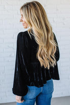Woman wearing a black velvet top and blue jeans against a white brick wall.