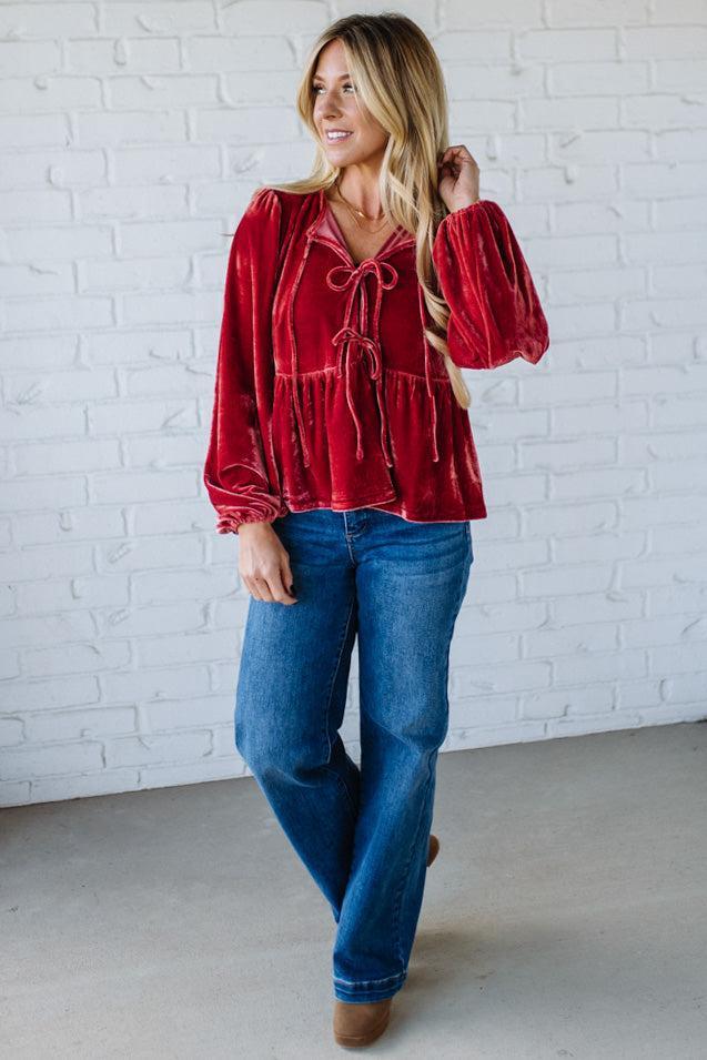 Woman wearing a red blouse and blue jeans against a white brick wall.