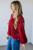 Woman wearing a red velvet top and blue jeans against a white brick wall.