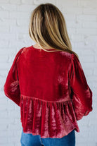Red velvet blouse with long sleeves worn by a person against a white brick wall.