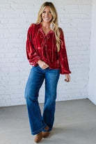 Woman wearing a red tie-dye blouse and blue jeans against a white brick wall.