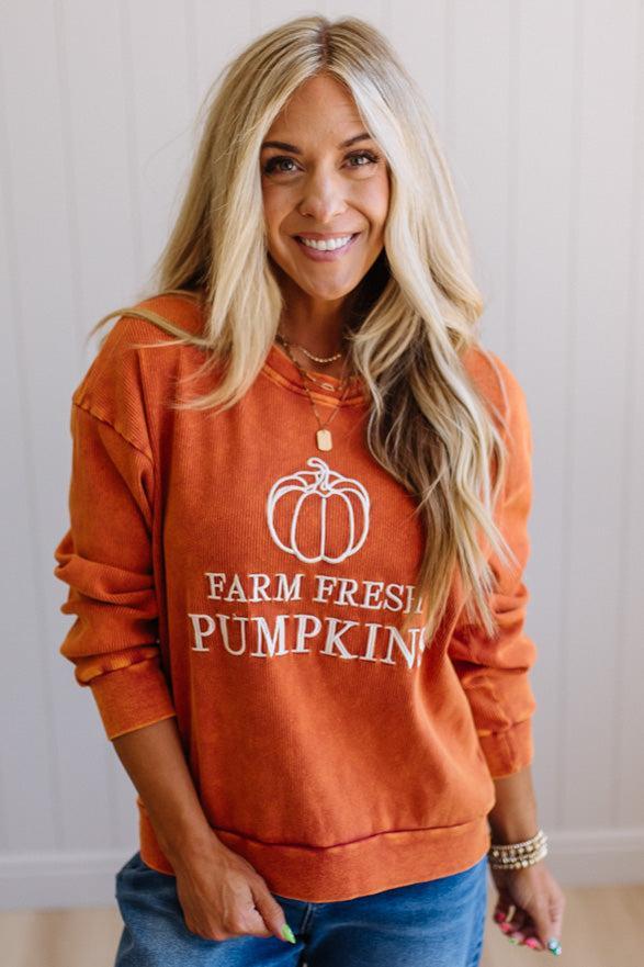 Blonde woman wearing a rust orange colored corduroy sweatshirt with white embroidered pumpkin and text: FARM FRESH PUMPKINS
