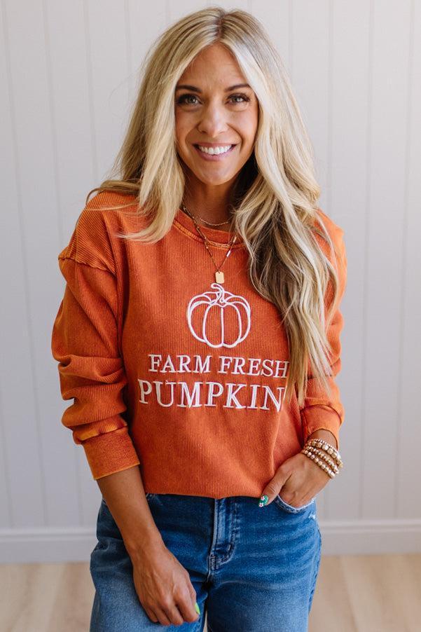 Blonde woman wearing a rust orange colored corduroy sweatshirt with white embroidered pumpkin and text: FARM FRESH PUMPKINS