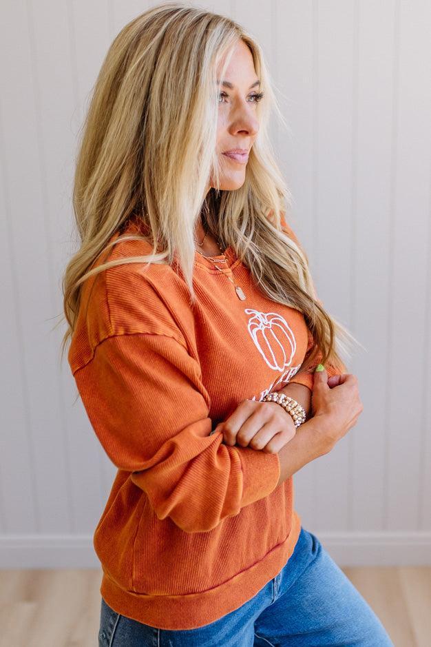 Blonde woman wearing a rust orange colored corduroy sweatshirt with white embroidered pumpkin and text: FARM FRESH PUMPKINS