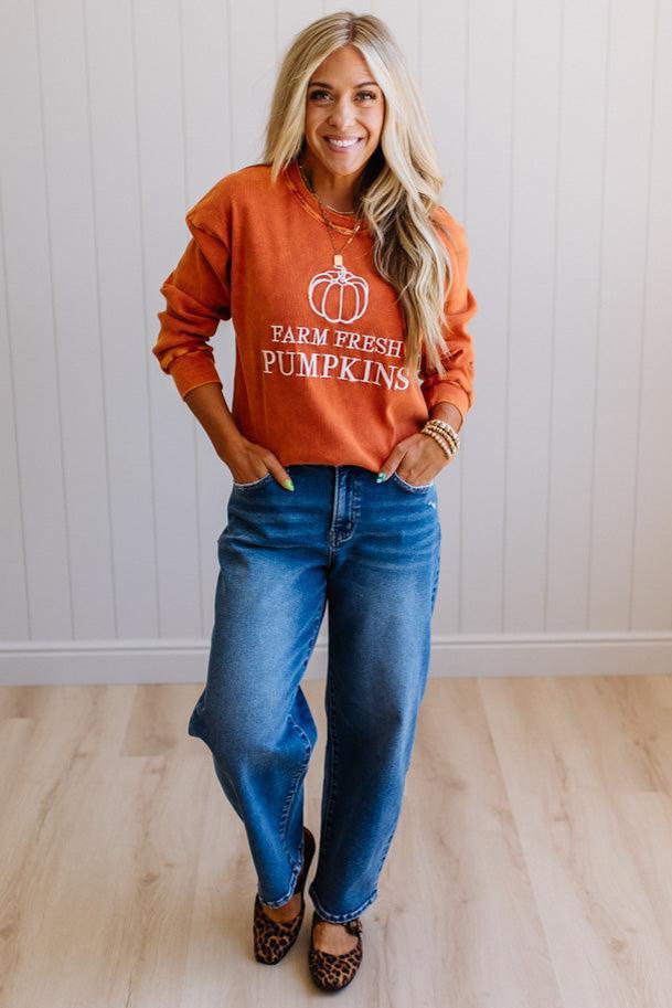 Blonde woman wearing a rust orange colored corduroy sweatshirt with white embroidered pumpkin and text: FARM FRESH PUMPKINS