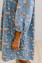 A close-up of a person wearing a blue floral dress with long sleeves, tie cuffs, and a side pocket. The dress has a vintage-style floral print in shades of blue, pink, and beige.
