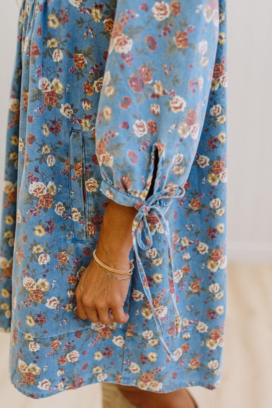 A close-up of a person wearing a blue floral dress with long sleeves, tie cuffs, and a side pocket. The dress has a vintage-style floral print in shades of blue, pink, and beige.