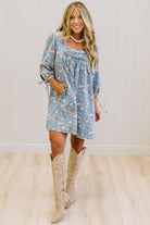 A woman modeling a light blue floral long-sleeve mini dress with tie details on the sleeves, paired with beige cowboy boots, standing in front of a white paneled wall and light wood floor.