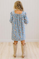 A woman viewed from behind wearing a light blue floral long-sleeve dress and beige cowboy boots, standing on a light wood floor against a white paneled wall.