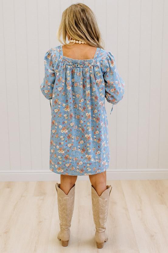 A woman viewed from behind wearing a light blue floral long-sleeve dress and beige cowboy boots, standing on a light wood floor against a white paneled wall.