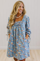 A woman wearing a light blue floral long-sleeve dress with a smocked bodice and tie details on the sleeves, standing against a white paneled wall.