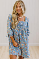A woman wearing a light blue floral long-sleeve dress with a smocked chest and tie detail on the sleeve, standing against a white paneled wall.