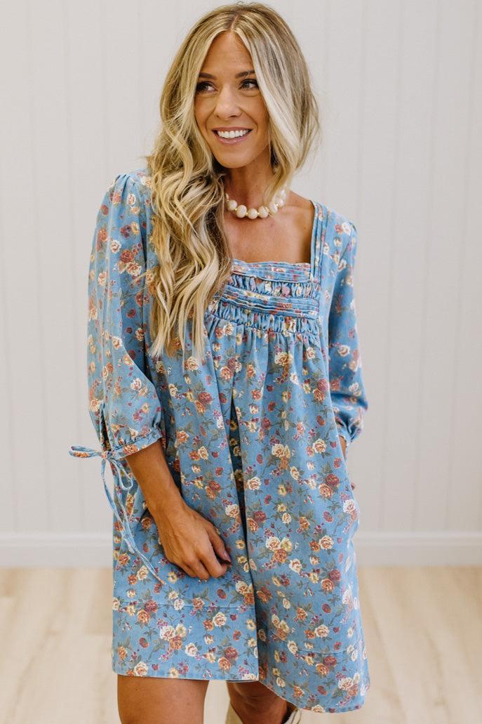 A woman wearing a light blue floral long-sleeve dress with a smocked chest and tie detail on the sleeve, standing against a white paneled wall.