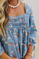 A close-up of a woman wearing a blue floral long-sleeve top with a smocked bodice and tie details at the cuffs, paired with a pearl necklace.