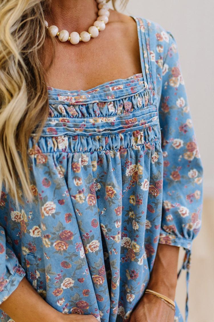 A close-up of a woman wearing a blue floral long-sleeve top with a smocked bodice and tie details at the cuffs, paired with a pearl necklace.