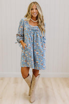 A woman modeling a light blue floral long-sleeve dress with a smocked neckline and tie details on the sleeves, paired with beige knee-high boots against a white paneled wall and light wood floor.