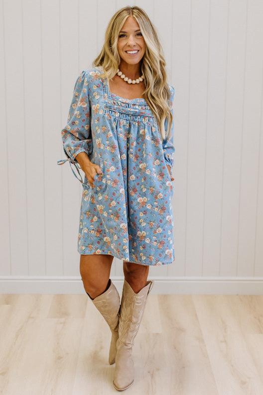 A woman modeling a light blue floral long-sleeve dress with a smocked neckline and tie details on the sleeves, paired with beige knee-high boots against a white paneled wall and light wood floor.
