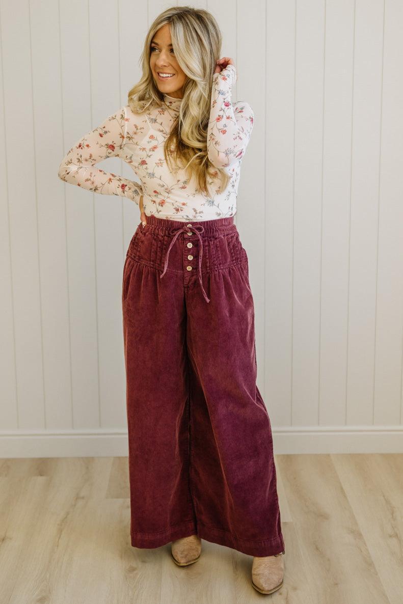 Woman wearing a floral blouse and burgundy wide-leg pants against a white paneled wall.