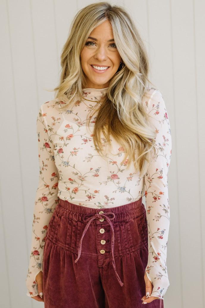 Woman wearing a floral long-sleeve top and burgundy pants against a light wooden panel background