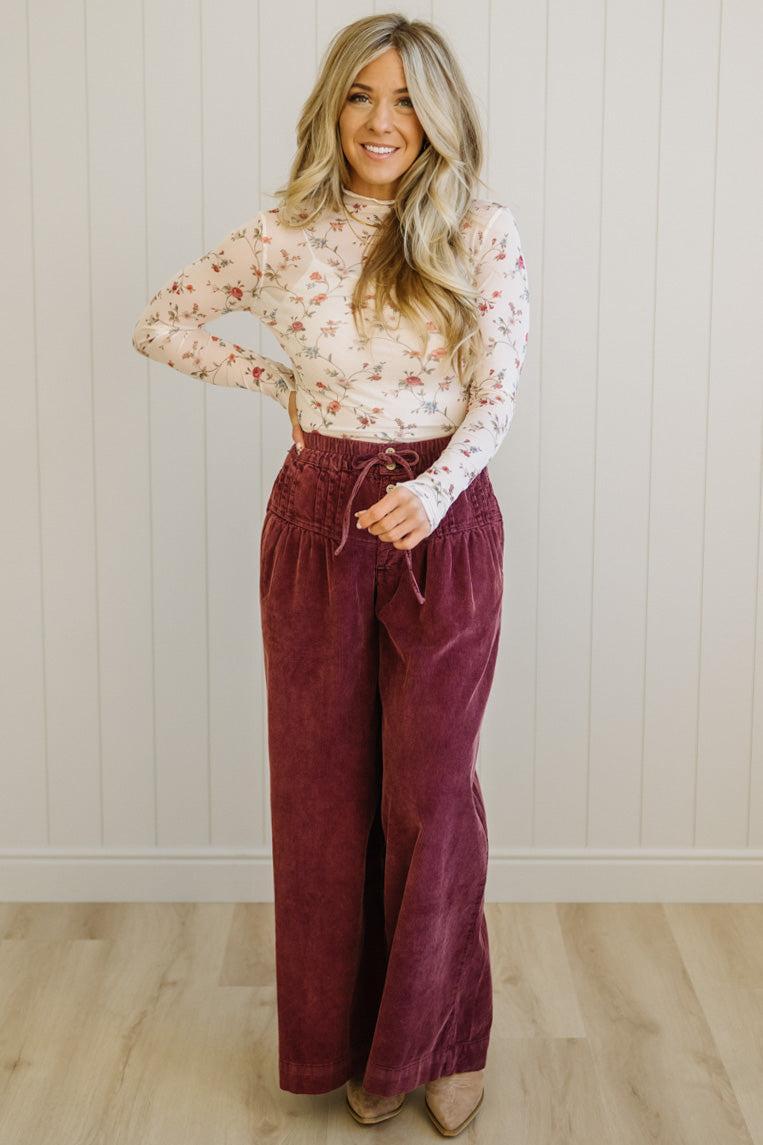 Woman wearing a floral blouse and burgundy pants against a white paneled wall.