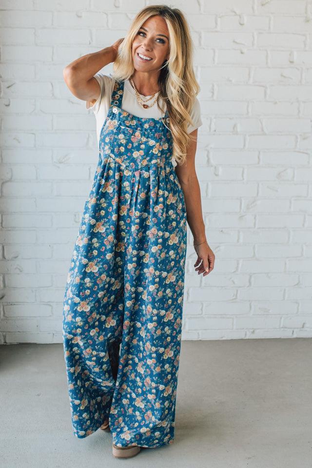 Woman wearing blue floral overalls against a white brick wall.
