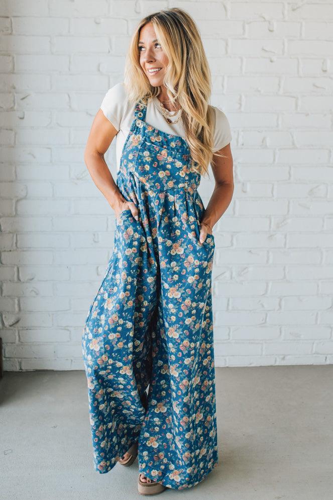 Woman wearing a blue floral jumpsuit against a white brick wall.