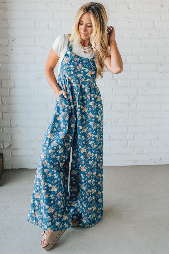 Woman wearing a blue floral jumpsuit against a white brick wall.