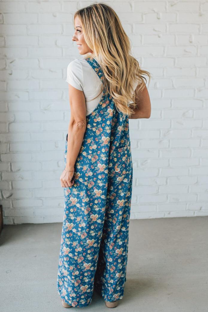 Woman wearing a blue floral jumpsuit against a white brick wall.