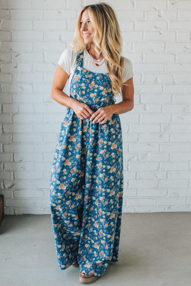 Woman wearing blue floral overalls standing against a white brick wall.
