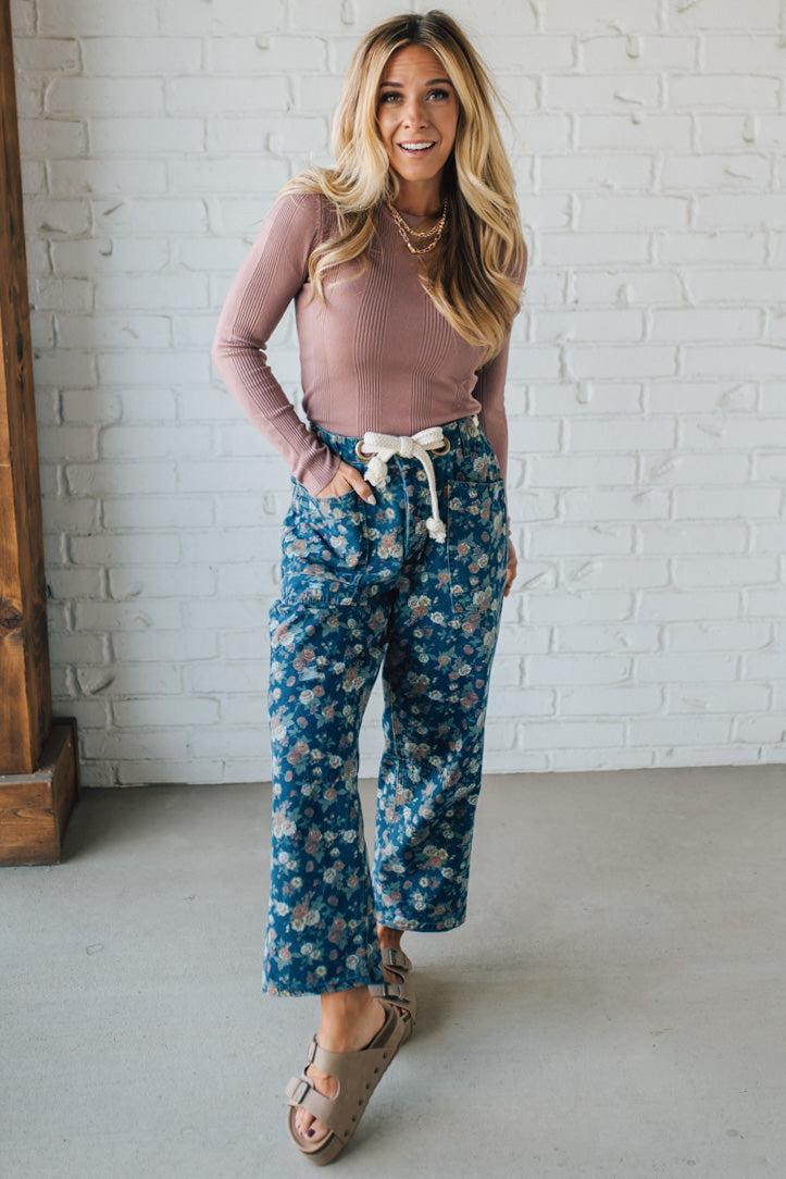 Woman wearing a pink long-sleeve top and blue floral pants against a white brick wall.