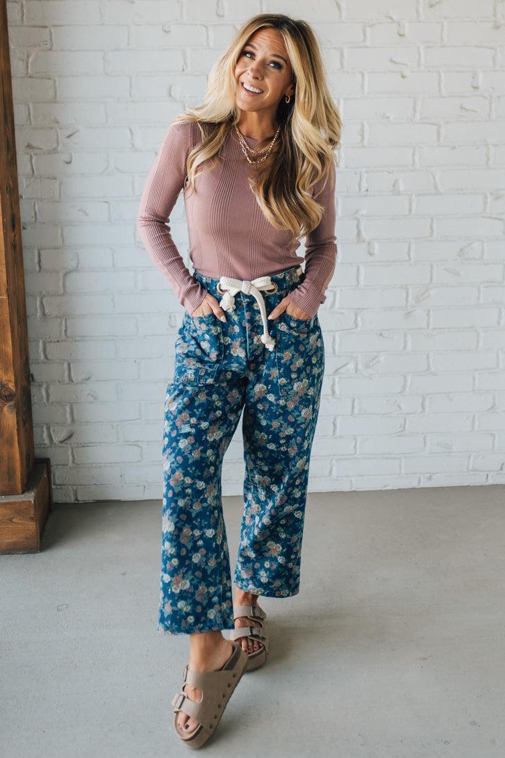 Woman wearing a pink top and blue floral pants against a white brick wall.