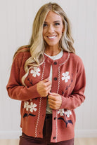 A smiling blonde woman wearing a rust-colored knit cardigan with white floral embroidery and cream stitched trim, layered over a white top and dark pants.