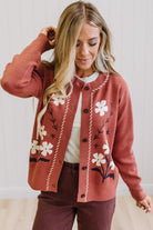 A woman wearing a rust-colored cardigan with white floral embroidery and cream rope trim, styled over a white top and dark brown pants.