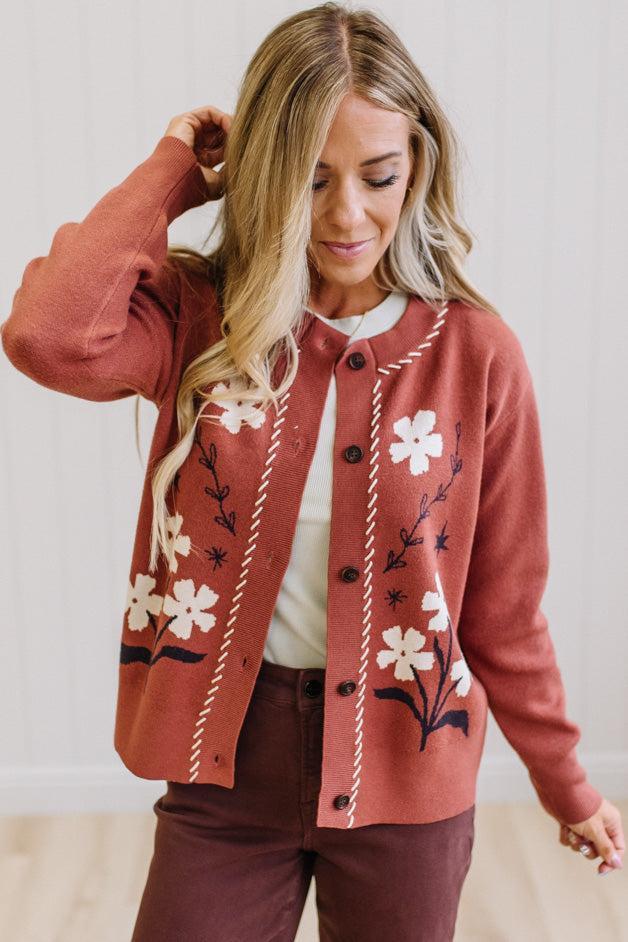 A woman wearing a rust-colored cardigan with white floral embroidery and cream rope trim, styled over a white top and dark brown pants.